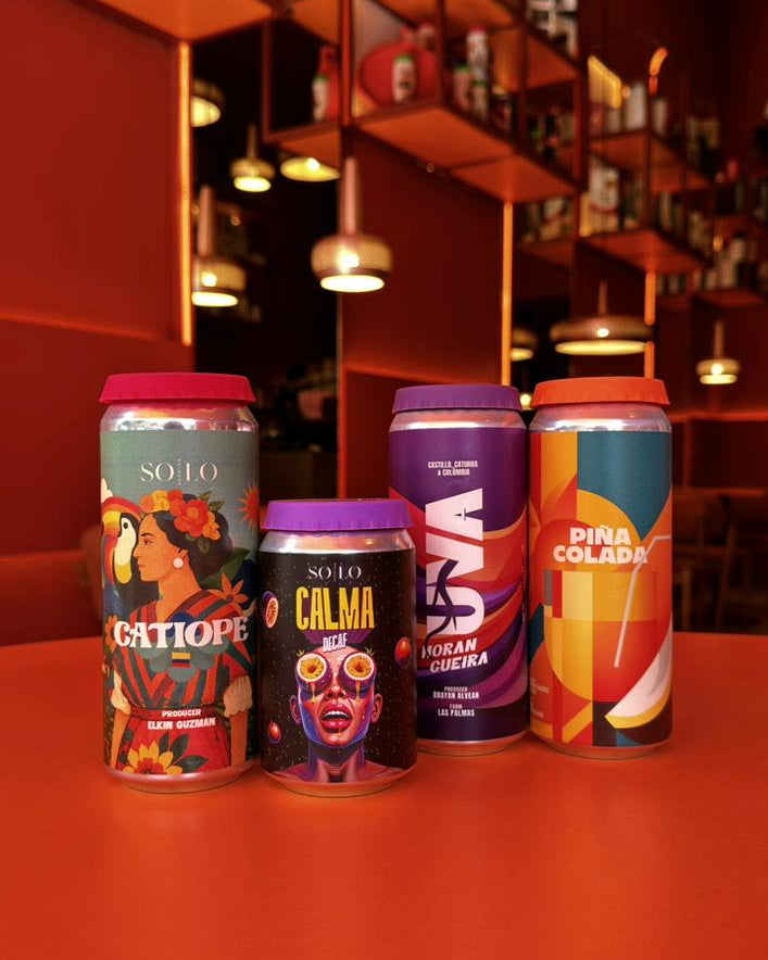 Four colorful beer cans with artistic designs on a red surface, with a bar setting in the background.